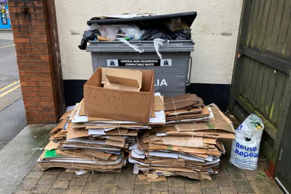 A photo of a large business waste container overflowing with rubbish and a large pile of cardboard in front